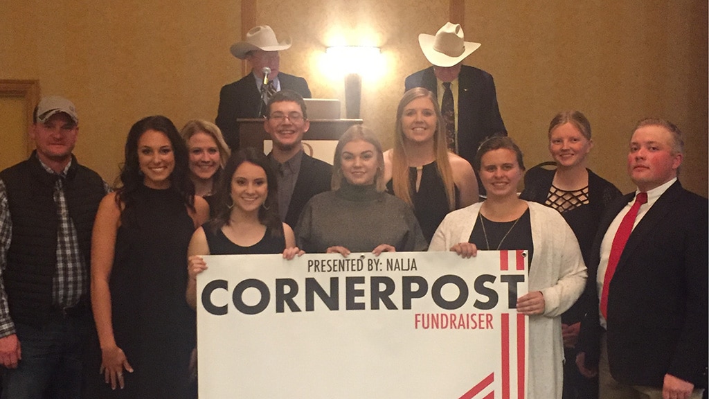 A group of people pose indoors, some smiling, with two men in cowboy hats at a podium behind them. The front row holds a large sign reading Cornerpost Fundraiser Presented by: NALJA.