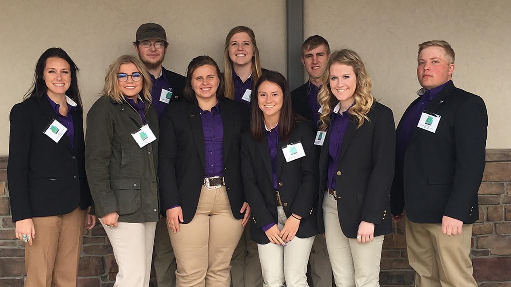 A group of nine young adults, dressed in business casual attire with nametags, stand together smiling in front of a beige wall with stone accents.