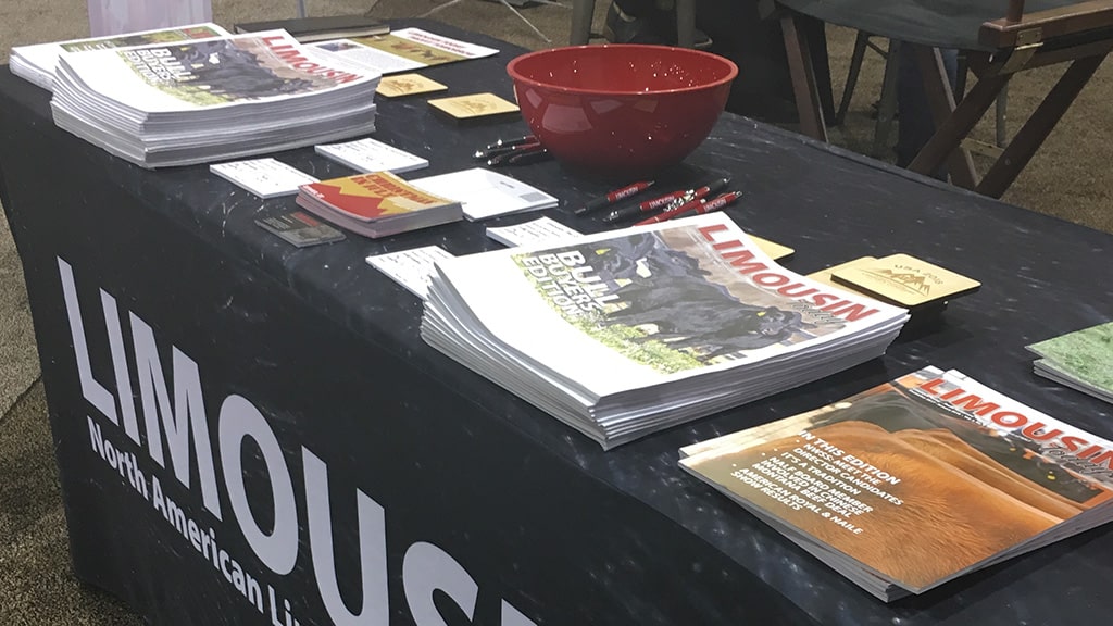 A table displays stacks of Limousin magazines, business cards, pens, notepads, and a red bowl. The table cover reads “North American Limousin.” The arrangement suggests a promotional or informational booth.