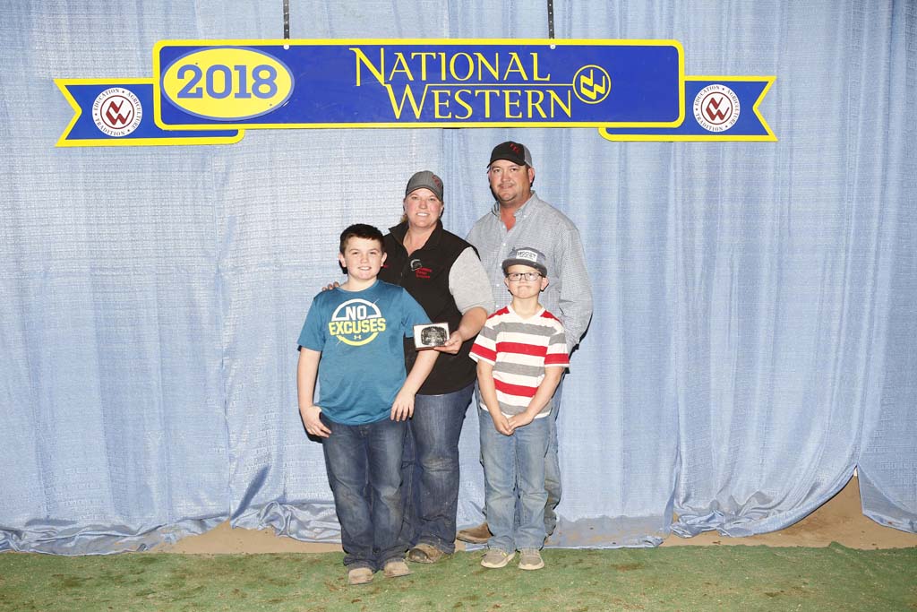 A family of four poses in front of a blue curtain and a yellow 2018 National Western sign. Two adults stand behind two boys; one holds an award. All are smiling and dressed casually.