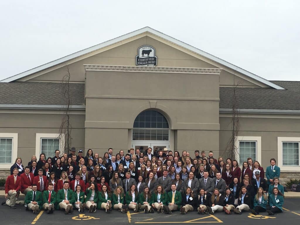 A large group of people, many wearing blazers in green, red, blue, or maroon, pose for a group photo in front of a building with a sign that reads Certified Angus Beef.
