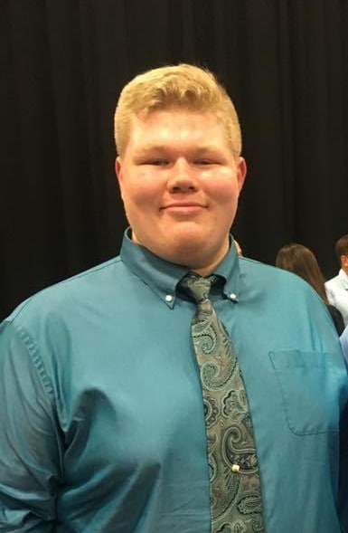 A young man with short blond hair is wearing a teal dress shirt and a paisley-patterned tie, standing indoors in front of a dark background.