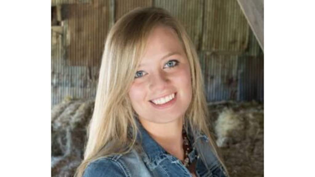 A young woman with long blonde hair smiles at the camera. She is wearing a denim jacket and a necklace, standing in front of a rustic, corrugated metal and wood background.