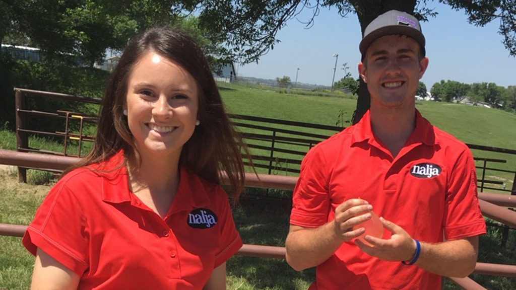 Two young adults wearing matching red shirts with naija logos are standing outdoors by a metal fence, smiling at the camera on a sunny day. There are green trees and grass in the background.