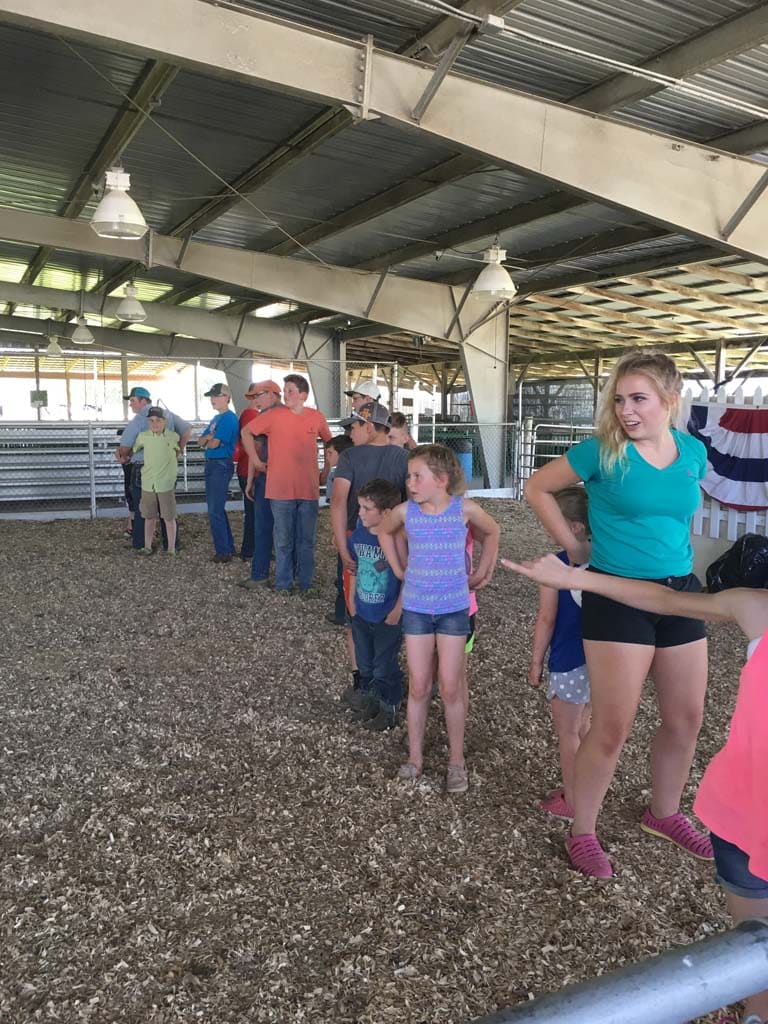 A group of children and adults stand in two lines inside a barn or covered arena, on a wood-chip floor, possibly waiting for an event or activity at a fair or livestock show.