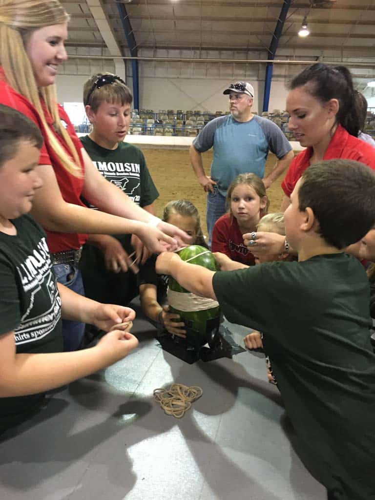 A group of children and two adults gather around a table indoors, placing rubber bands on a watermelon. Several rubber bands and the watermelon are on the table, and the scene appears lively and focused.