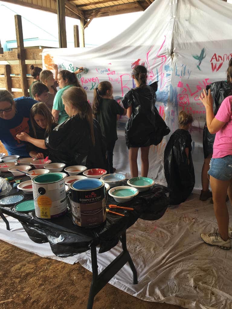 Children wearing garbage bags as smocks paint on a white tent wall, supervised by an adult. Various paint cans and bowls are arranged on a table in the foreground, and the scene is set in an open, covered space.