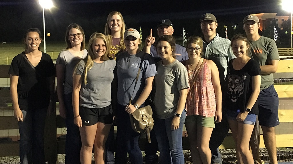 A group of eleven people, mostly young adults, smile for a photo at night outdoors near a wooden fence, with lights and a field visible in the background.