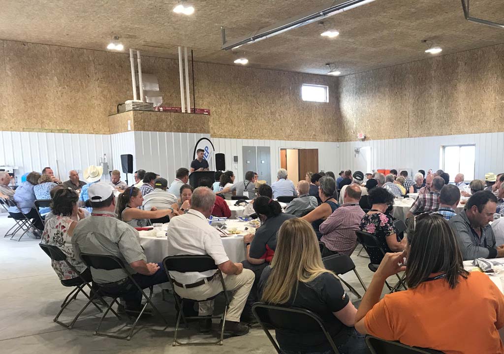 A large group of people sit at round tables in a spacious, well-lit indoor hall, listening to a speaker standing at a podium in the front of the room. The atmosphere appears to be that of a community event or gathering.