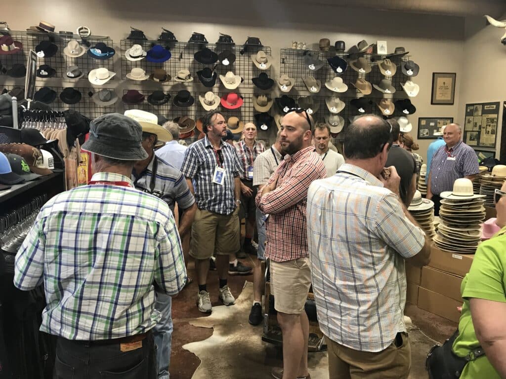 A group of men, mostly wearing plaid shirts and hats, gather in a store filled with various hats displayed on shelves and racks. The room is busy, and hats cover the walls in different styles and colors.