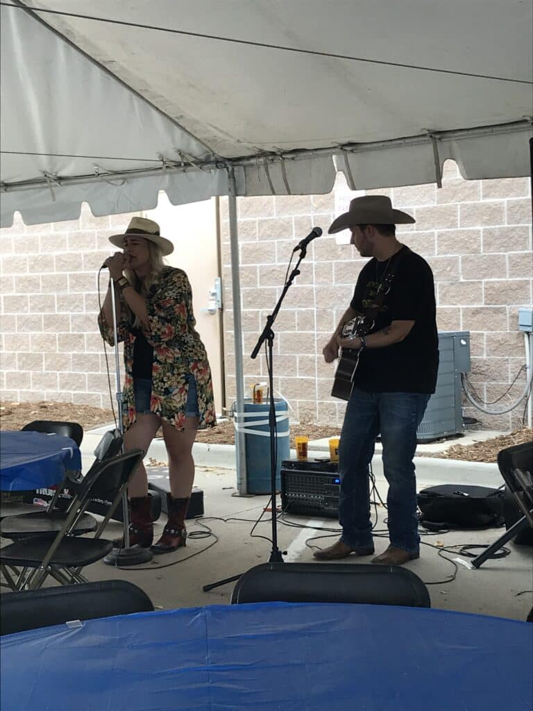 A woman sings into a microphone while a man plays acoustic guitar beside her. Both wear cowboy hats and stand under a tent, with empty chairs and tables in the foreground.