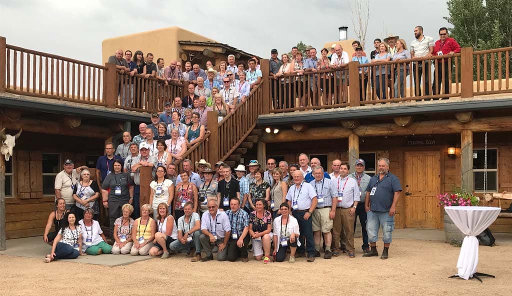 A large group of people pose together for a group photo outside a rustic, wooden lodge with two stories. Some stand on the balcony and stairs, while others are gathered in front of the building.