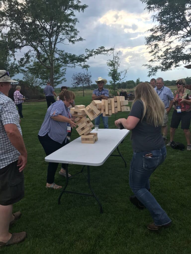 A group of people outdoors watch as two women play giant Jenga on a table; the wooden blocks are collapsing mid-game. Trees and grass surround the scene under a cloudy sky.