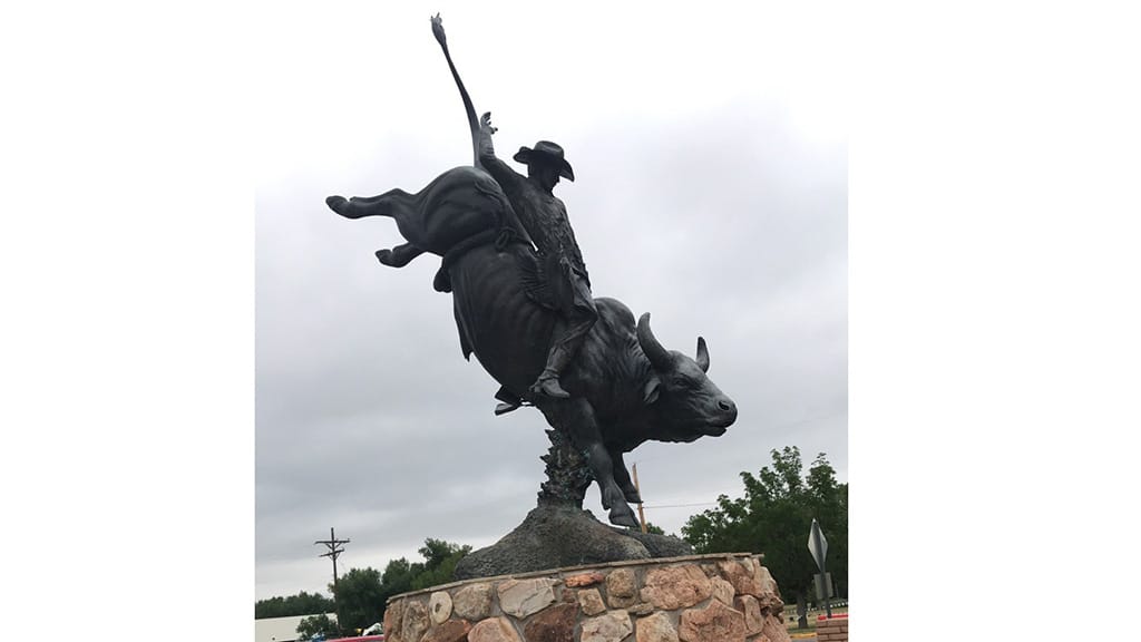 A bronze statue of a rodeo cowboy riding a bucking bull, with one arm raised, stands on a stone base outdoors against a cloudy sky. Trees and utility poles are visible in the background.