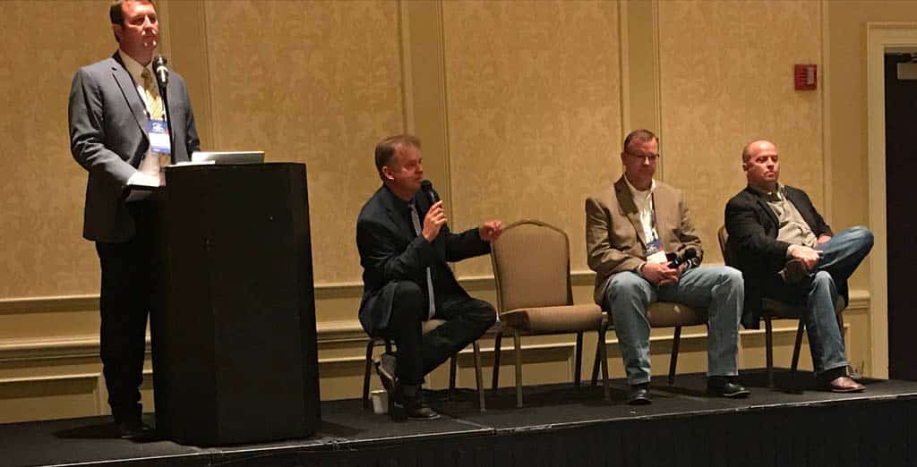 Four men are at the front of a room; one stands at a podium speaking, while three sit in chairs on stage, with one of the seated men holding a microphone. The setting appears to be a conference or panel discussion.