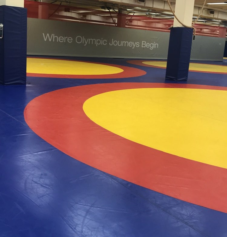 An empty wrestling gym with blue, red, and yellow mats. The wall in the background has the words Where Olympic Journeys Begin written on it.