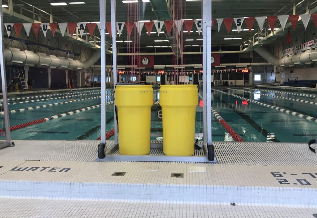 Two large yellow trash bins stand side by side at the edge of an indoor swimming pool, near lane dividers and diving boards, in a well-lit aquatic center.