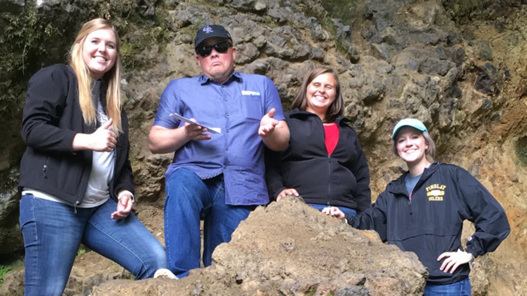 Four people pose in front of a rocky wall. Three women stand on either side of a man in sunglasses and a cap, who shrugs with both hands. All are smiling and appear to be enjoying a casual outdoor outing.