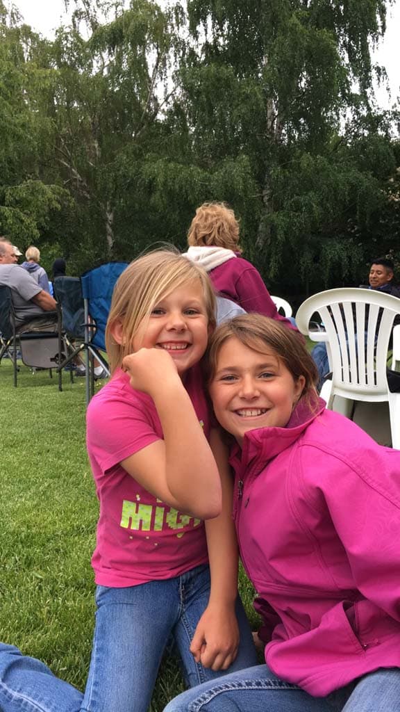 Two young girls sitting on grass, smiling and hugging each other, both wearing pink tops and jeans. People are sitting on chairs in the background with trees and greenery surrounding them.