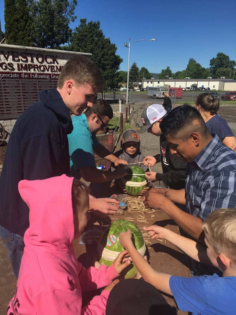 A group of people, including children and adults, gather around an outdoor table cutting open watermelons on a sunny day. Some wear hoodies and hats, and trees and buildings are visible in the background.