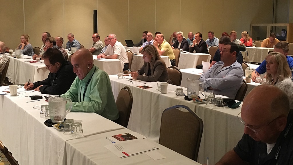 A group of people sit at tables in a conference room, listening attentively and taking notes during a meeting or seminar. The room is well-lit and organized, with water pitchers and papers on the tables.