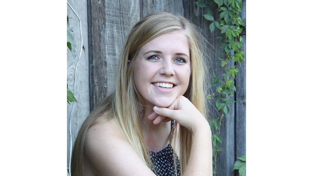 A young woman with long blonde hair smiles at the camera, resting her chin on her hand. She is sitting outdoors in front of a wooden fence with green vines.