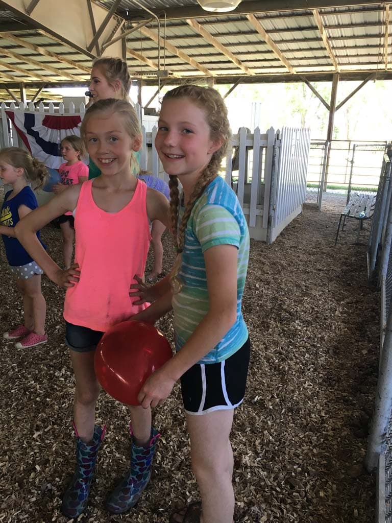 Two young girls stand smiling in a barn, one holding a red bowl. Both are wearing shorts and T-shirts, with one in pink and the other in blue stripes. Other kids and a white fence are visible in the background.