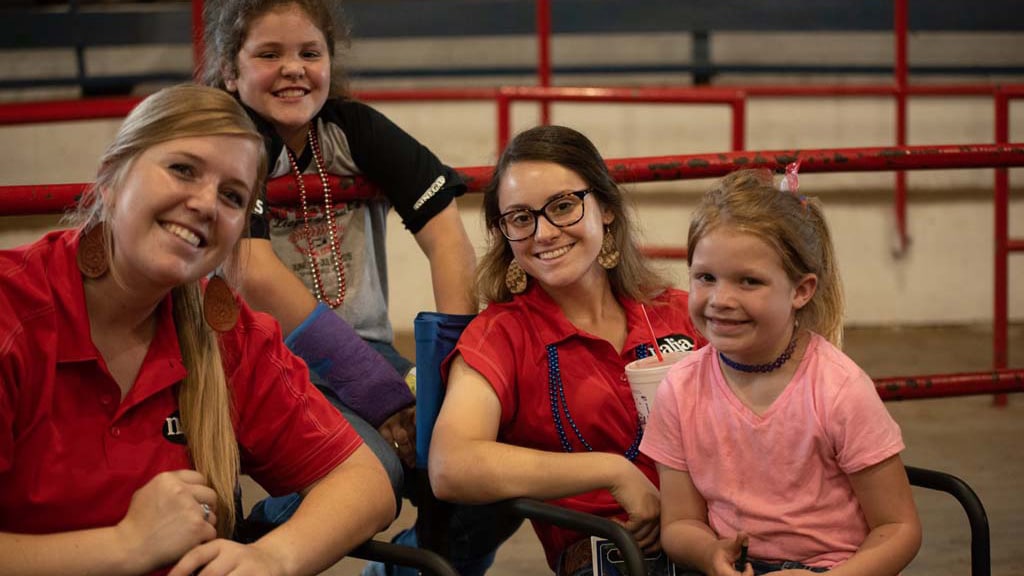 Four smiling girls, two in red shirts seated in front and two behind a metal railing, pose together indoors. One girl has a purple cast on her arm. The background is slightly blurred.