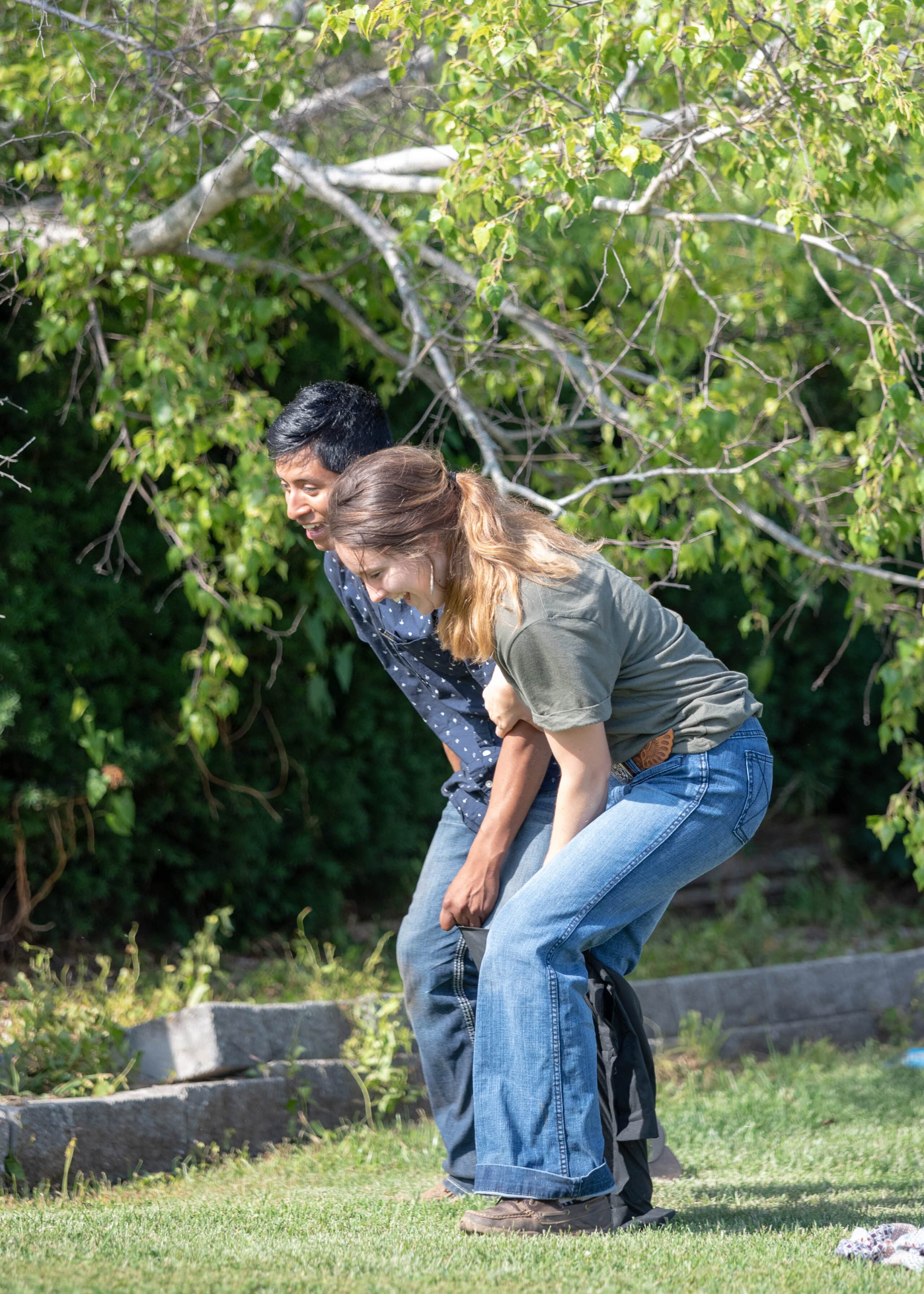 Two people are outdoors on grass, both bending forward and holding onto a bag. Trees and sunlight are in the background, and they appear to be engaged in a playful activity or teamwork task.