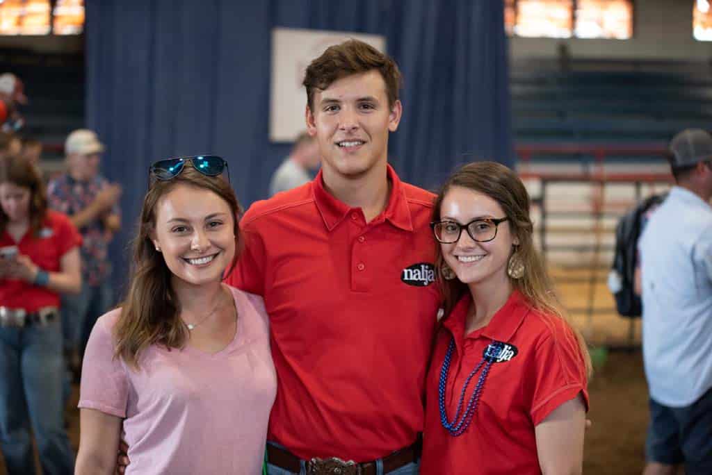 Three young adults smile for a photo at an indoor event. Two women stand on either side of a man in a red shirt. There are people and blue curtains in the background.