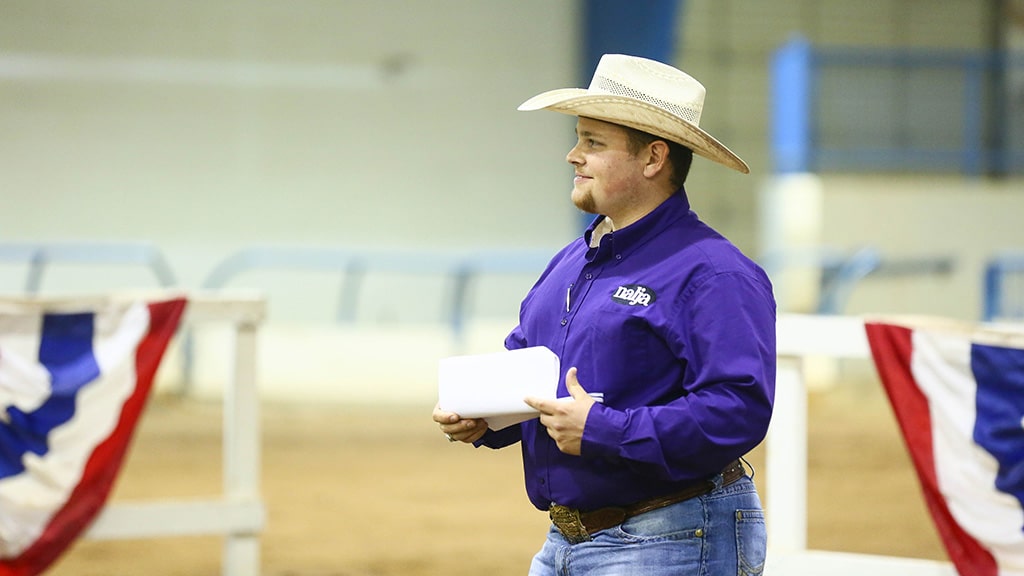 A man in a cowboy hat and purple shirt holds papers while standing in an indoor arena decorated with red, white, and blue bunting.