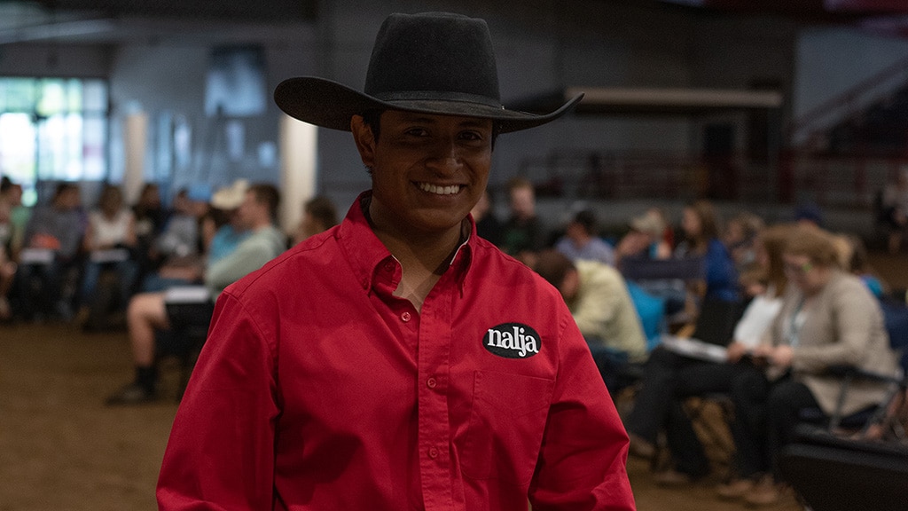 A person wearing a black cowboy hat and a red shirt with a najs logo smiles at the camera. In the background, people sit in rows at an indoor event.