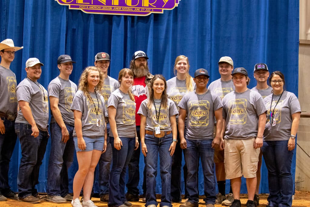 A group of people wearing matching gray event t-shirts pose and smile together in front of a blue curtain with a yellow and purple sign overhead. Most are wearing hats and badges.