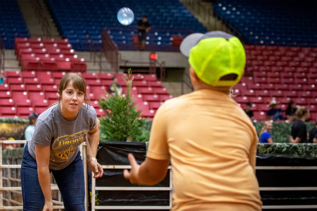 A woman and a boy in a neon cap play catch with a blue ball inside an arena with empty red and blue seats in the background. The woman is focused on the ball, while the boy prepares to catch it.