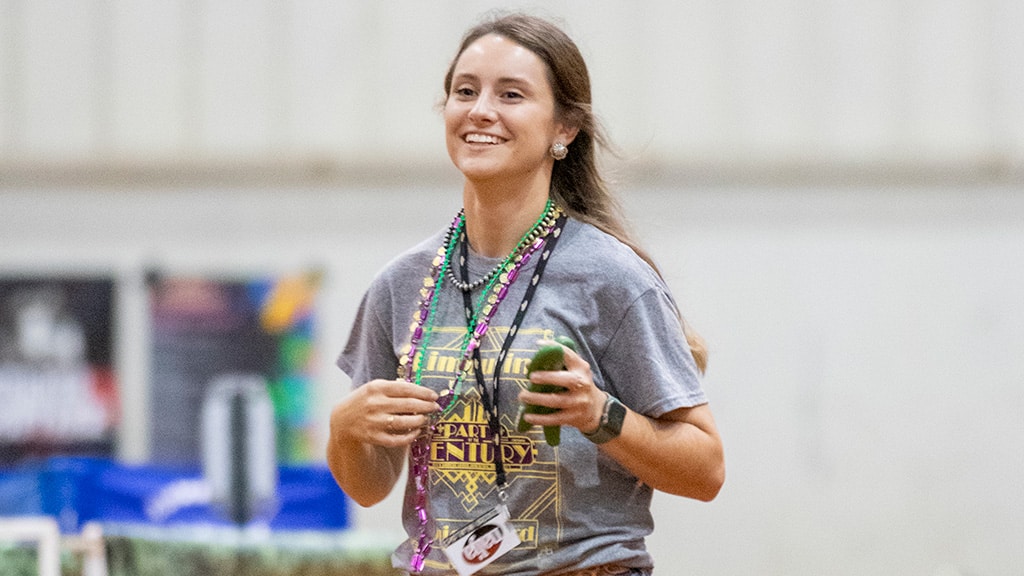 A smiling woman wearing a gray graphic t-shirt, colorful beaded necklaces, and a lanyard holds a green bell pepper and a small item while standing indoors.