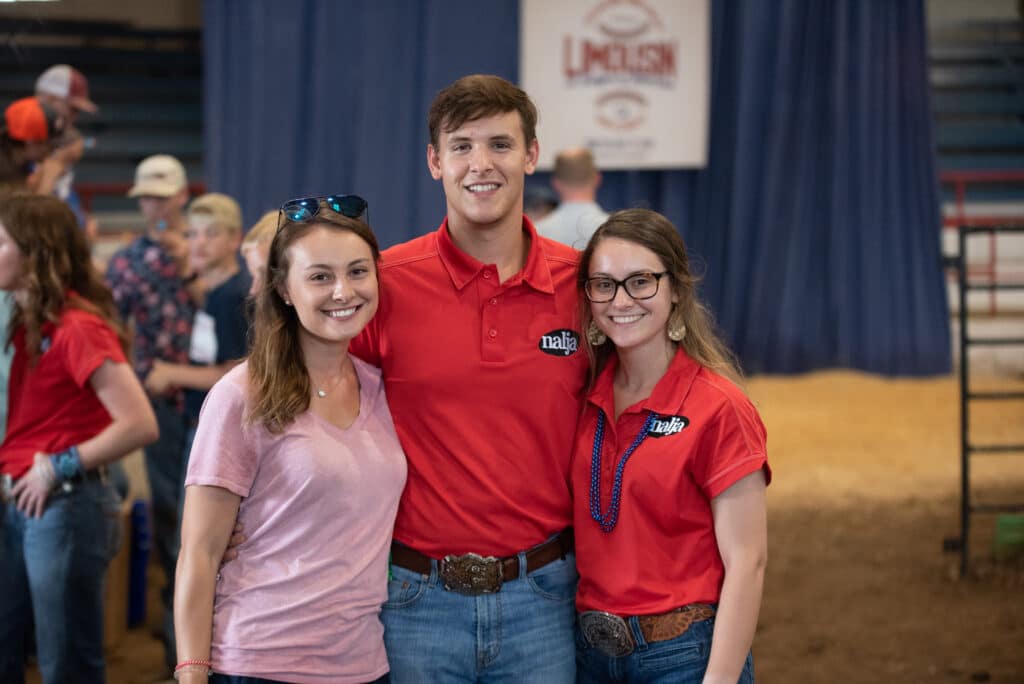 Three young adults smile at the camera in an indoor arena. Two women and one man stand together; the man and one woman wear matching red shirts with a logo, and the other woman wears a pink shirt. People and blue curtains are in the background.