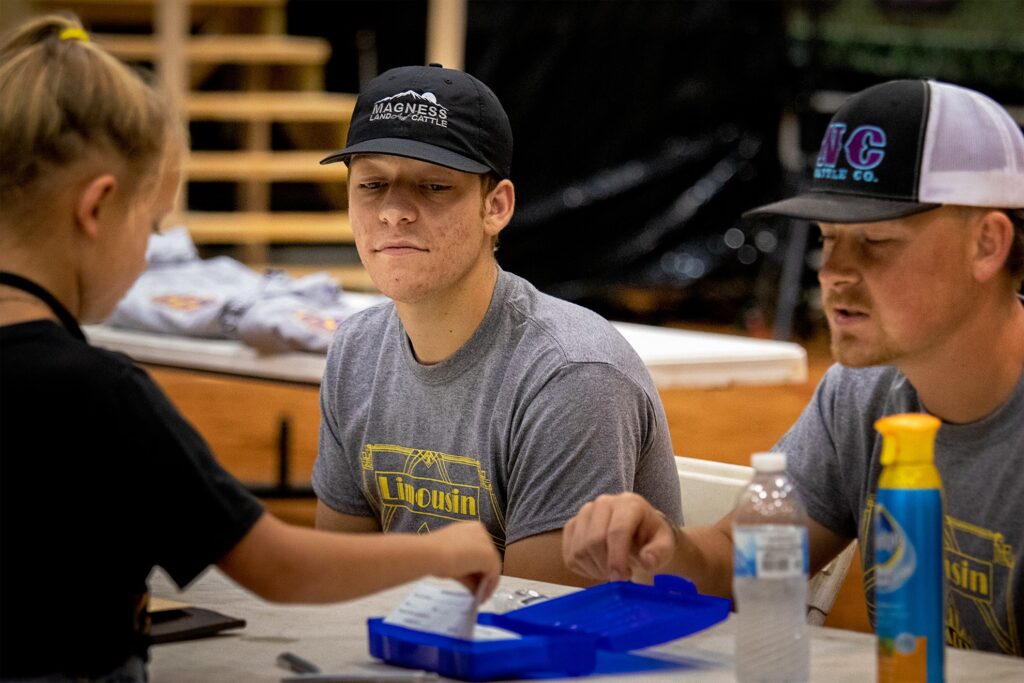 Two young men in gray shirts and baseball caps sit at a table, looking at papers handed to them by a young girl. A blue container, water bottle, and spray bottle are on the table. Indoors with wooden structures in background.