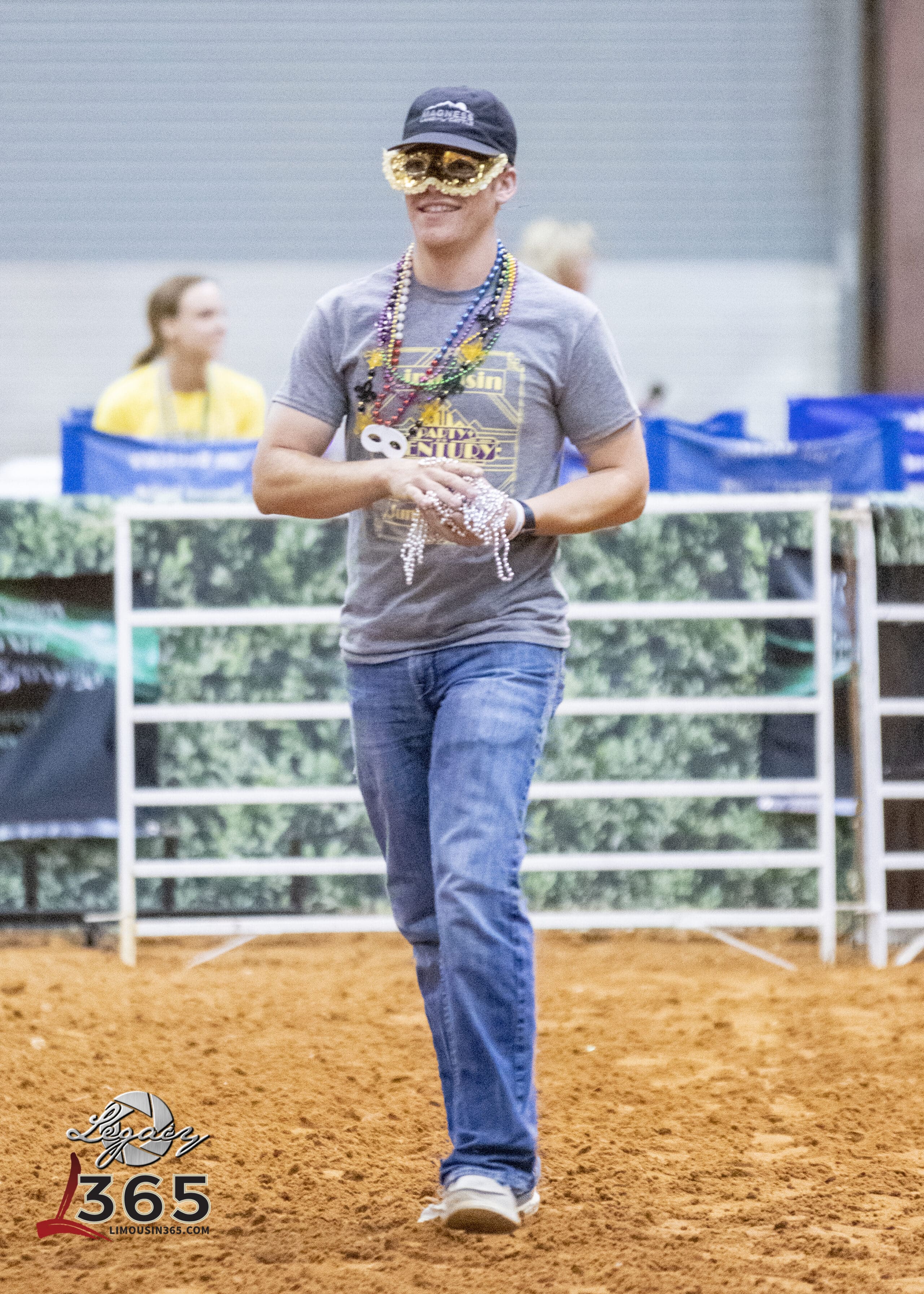 A person wearing a black cap, gold mask, and beaded necklaces walks on an indoor dirt arena, holding more beads. They are smiling and dressed casually in jeans and a T-shirt. The event logo Livestock 365 appears in the corner.