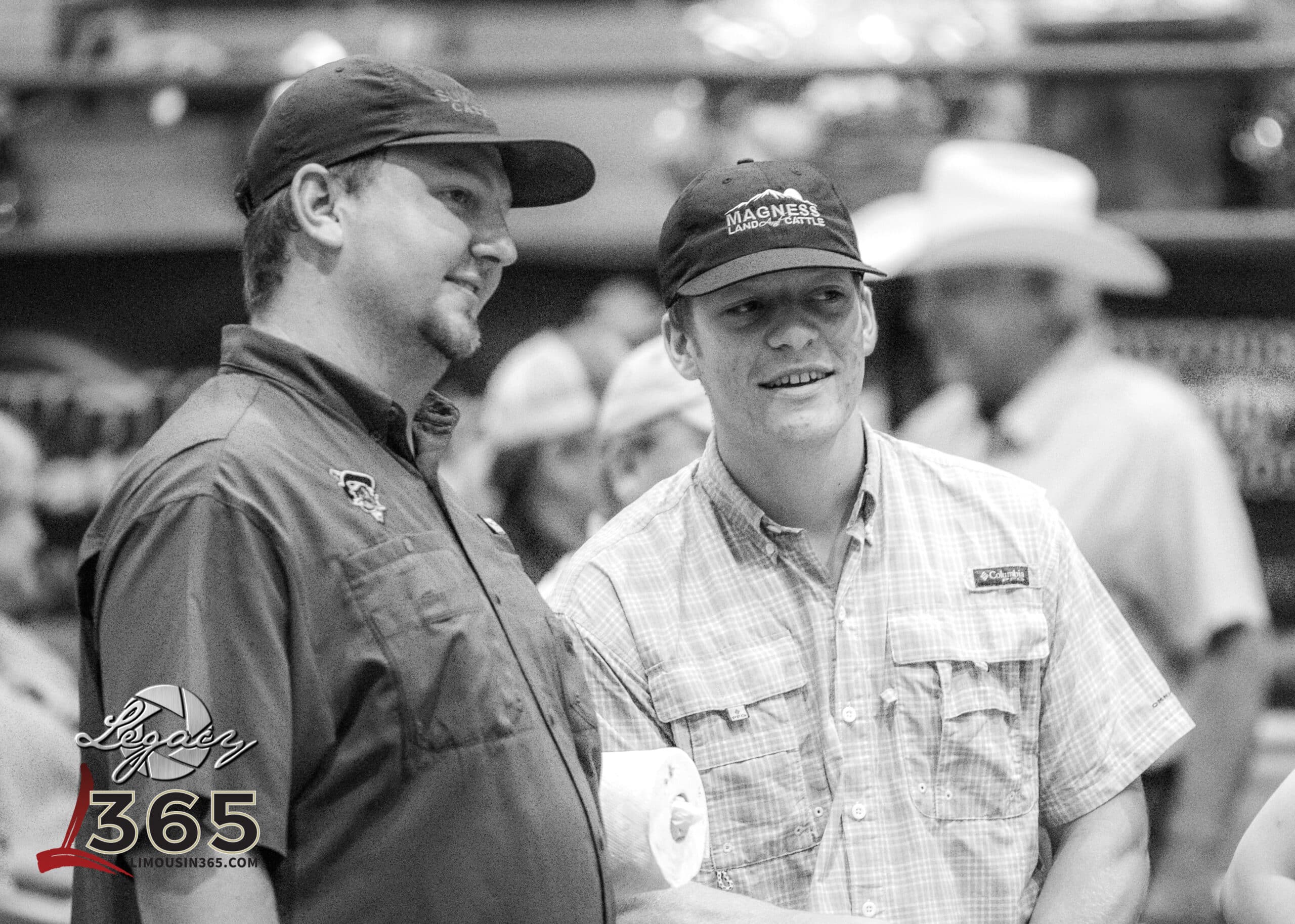 Two men in casual outdoor shirts and caps stand next to each other, smiling. One holds a piece of paper. Other people, some wearing hats, are blurred in the background. The image is black and white with the “Legacy 365” logo in the corner.