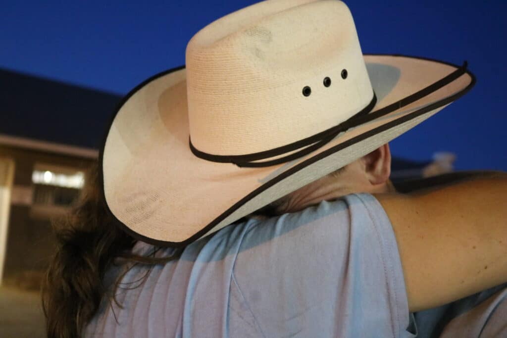 A person wearing a wide-brimmed cowboy hat embraces another person outside in the evening, their faces hidden from view. The background is dimly lit with a deep blue sky.