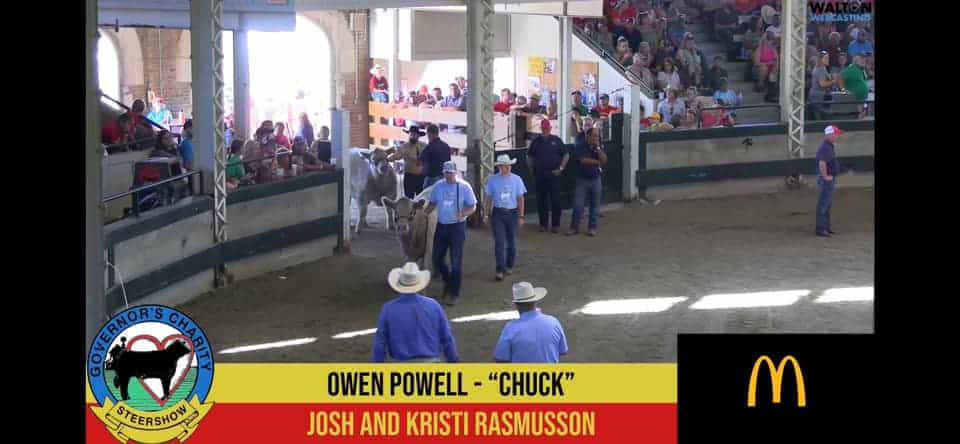 A livestock show arena with people in cowboy hats leading a steer. Spectators fill the stands. A banner reads “Governor’s Charity Steer Show.” Text on screen: “Owen Powell - ‘Chuck’, Josh and Kristi Rasmusson,” with McDonald’s logo.