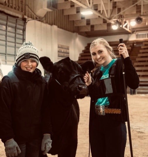 Two people smile while standing next to a black cow inside an indoor arena. One person wears a beanie and gloves, and the other holds a show stick, dressed in a belt and turquoise shirt. The background shows empty bleachers.