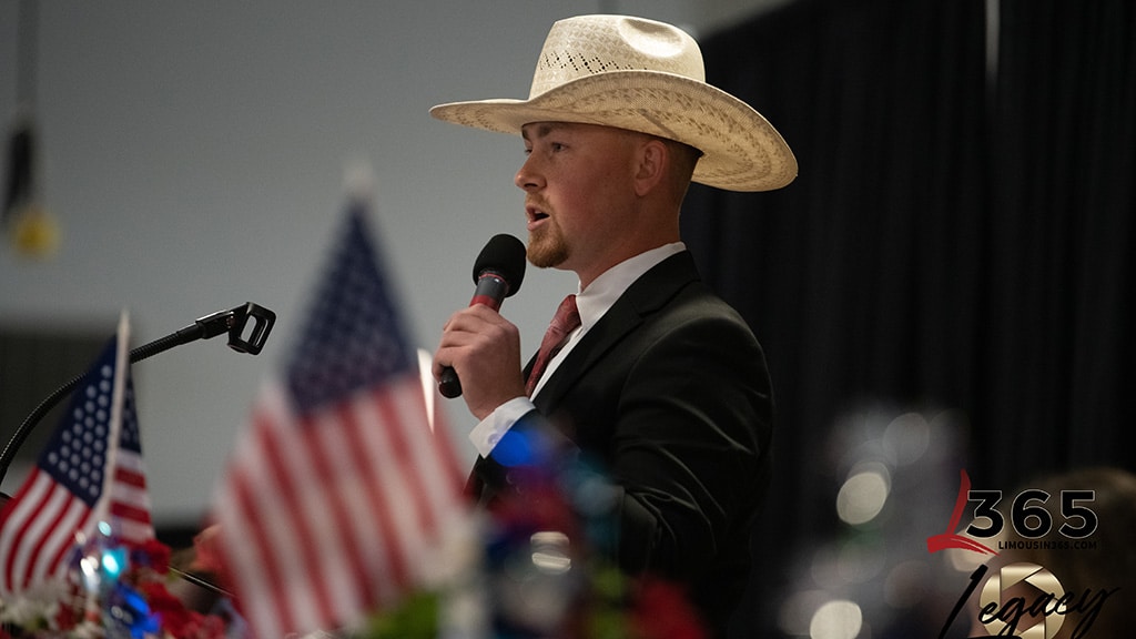 A man in a suit and cowboy hat speaks into a microphone at a podium, with small American flags and patriotic decorations in the foreground.