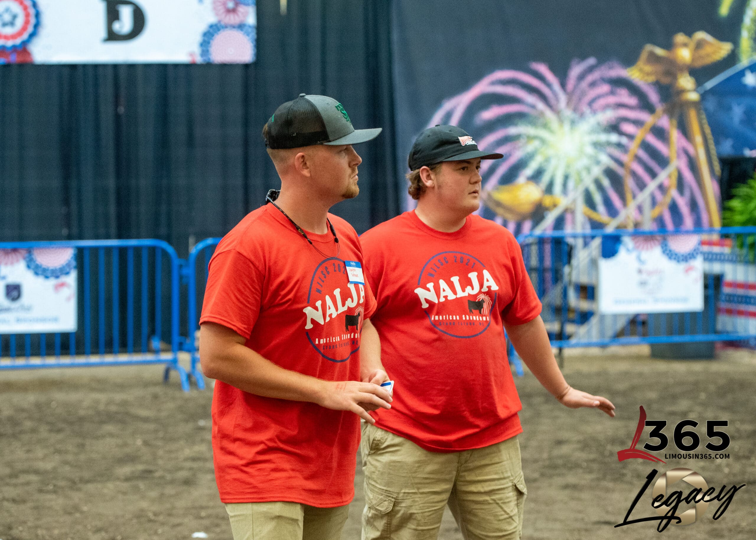 Two young men wearing red NALJA t-shirts and khaki shorts stand side by side in an indoor arena, looking ahead. Behind them are blue barriers and colorful decorations, including fireworks and patterned banners.