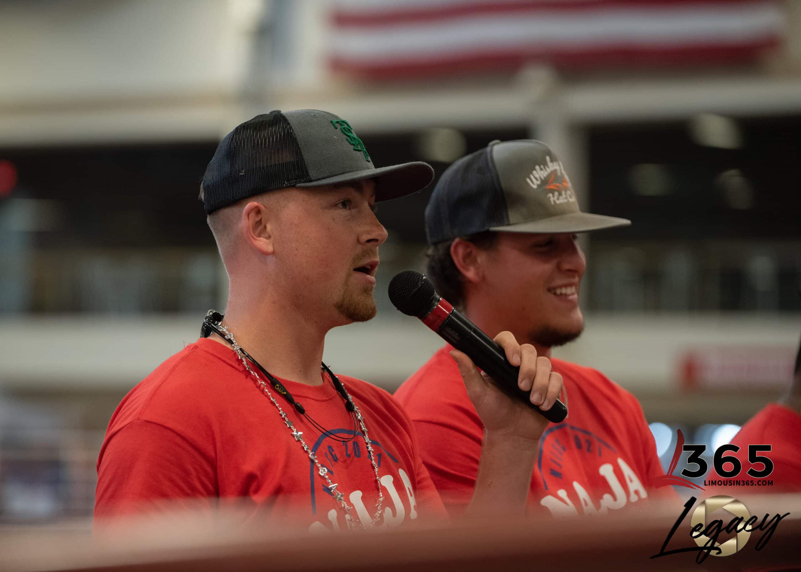 Two men in red shirts and baseball caps speak into a microphone indoors. One is holding the mic and wearing a beaded necklace; both are smiling. An American flag and blurred background are visible. 365 Legacy logo is at the bottom.