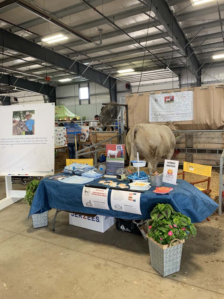 A decorated booth table with brochures, a donation box, and flowers is set up next to a cow in a barn-like indoor space, with signs and farming materials visible in the background.