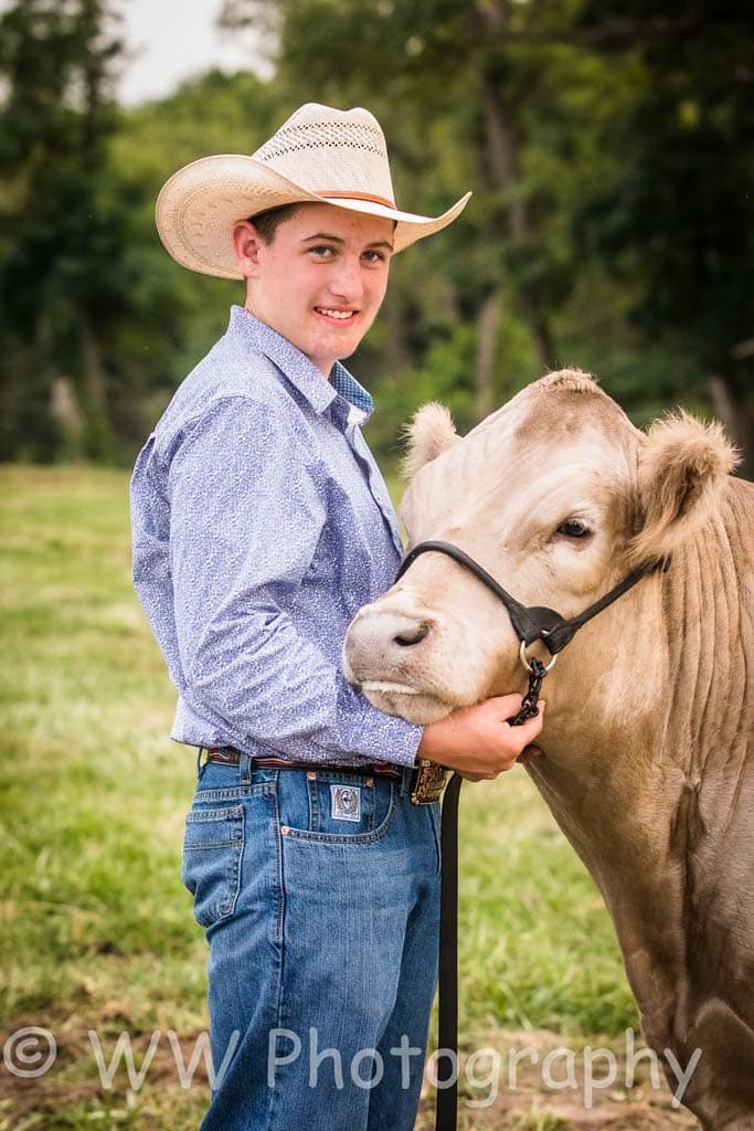 A young man in a cowboy hat and blue shirt smiles while holding a light brown cow by a halter in a grassy outdoor setting. WW Photography watermark is visible at the bottom.
