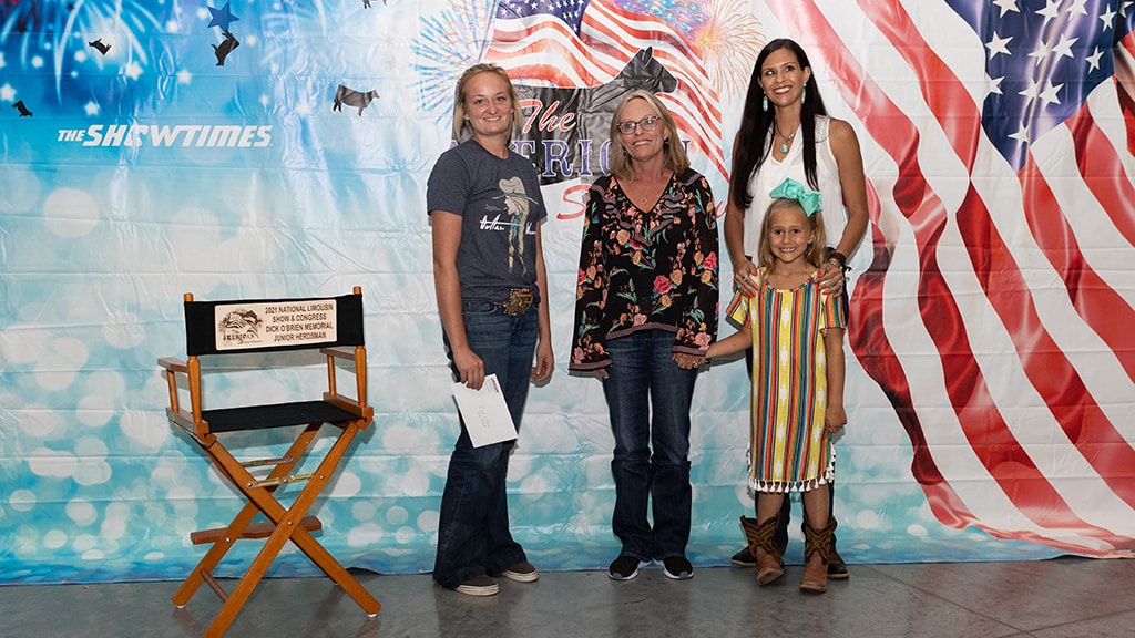 Four people, including a young girl in a striped dress and cowboy boots, pose smiling in front of a patriotic backdrop with an American flag. A director’s chair with a plaque sits on the left.