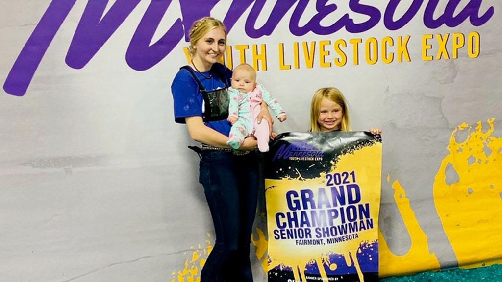 A smiling woman holding a baby stands next to a young girl who is holding a 2021 Grand Champion Senior Showman banner at the Minnesota Youth Livestock Expo in Fairmont, Minnesota.