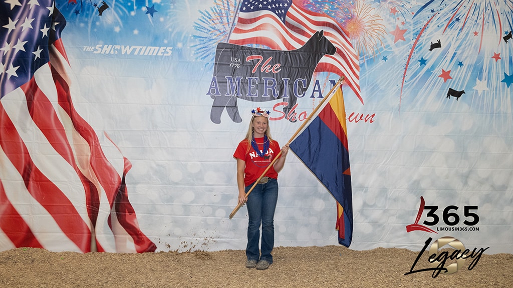A smiling woman in a patriotic outfit holds an Arizona state flag in front of a backdrop with American flags, a cow silhouette, and “The American Showman” text. Confetti decorates the ground.